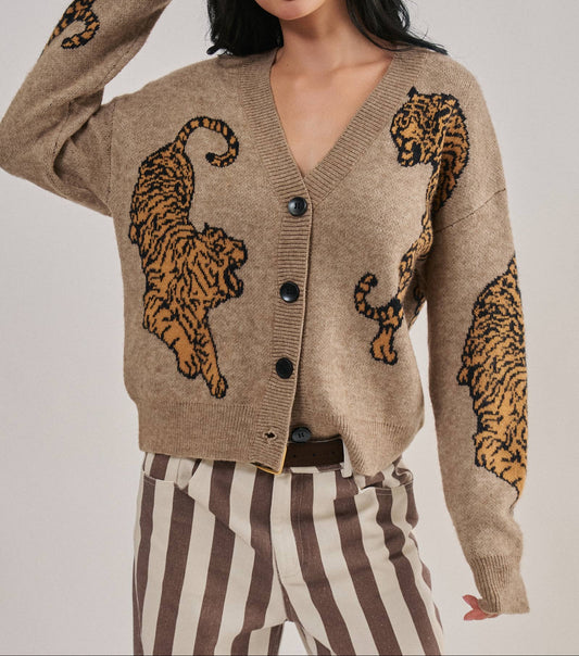 A woman wearing a beige knit cardigan with large tiger motifs on the front and sleeves, paired with brown and cream striped pants, against a light gray studio background.