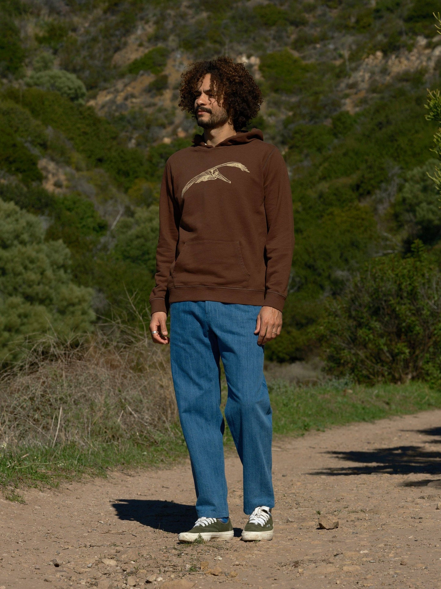 Person wearing a brown hoodie with a bird design outdoors on a dirt path.