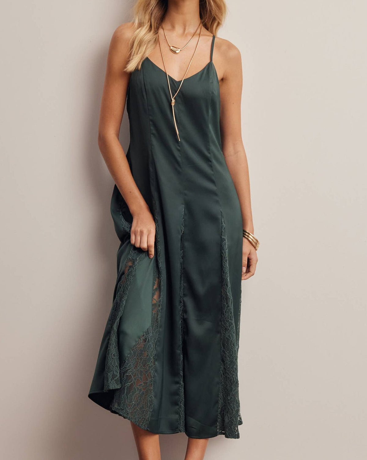 A woman models a dark green satin slip dress with lace trim and a matching long necklace against a neutral beige wall.