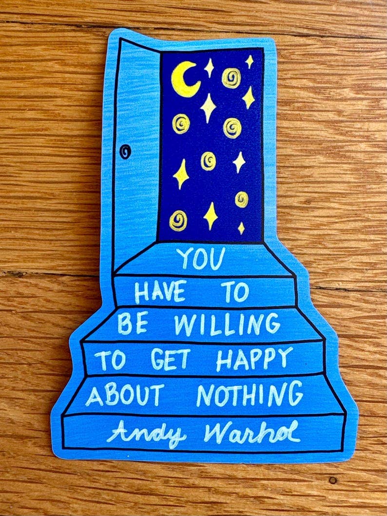 A hand-painted Andy Warhol-style blue door-shaped sign with yellow stars and moons and white text reading 'YOU HAVE TO BE WILLING TO GET HAPPY ABOUT NOTHING Andy Warhol' on a wooden surface.