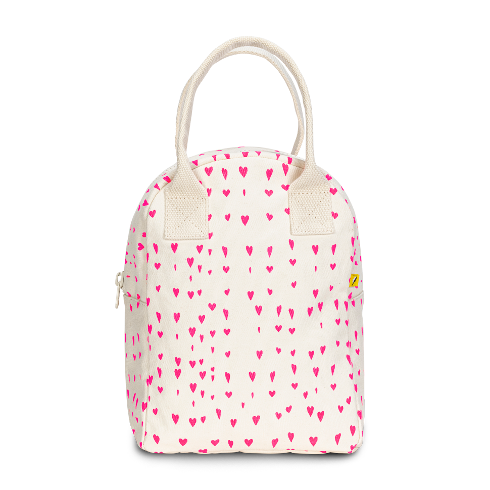 An off-white canvas lunch bag with a repeating pattern of small pink hearts, featuring cream-colored rope handles and a small yellow brand tag on the right side.