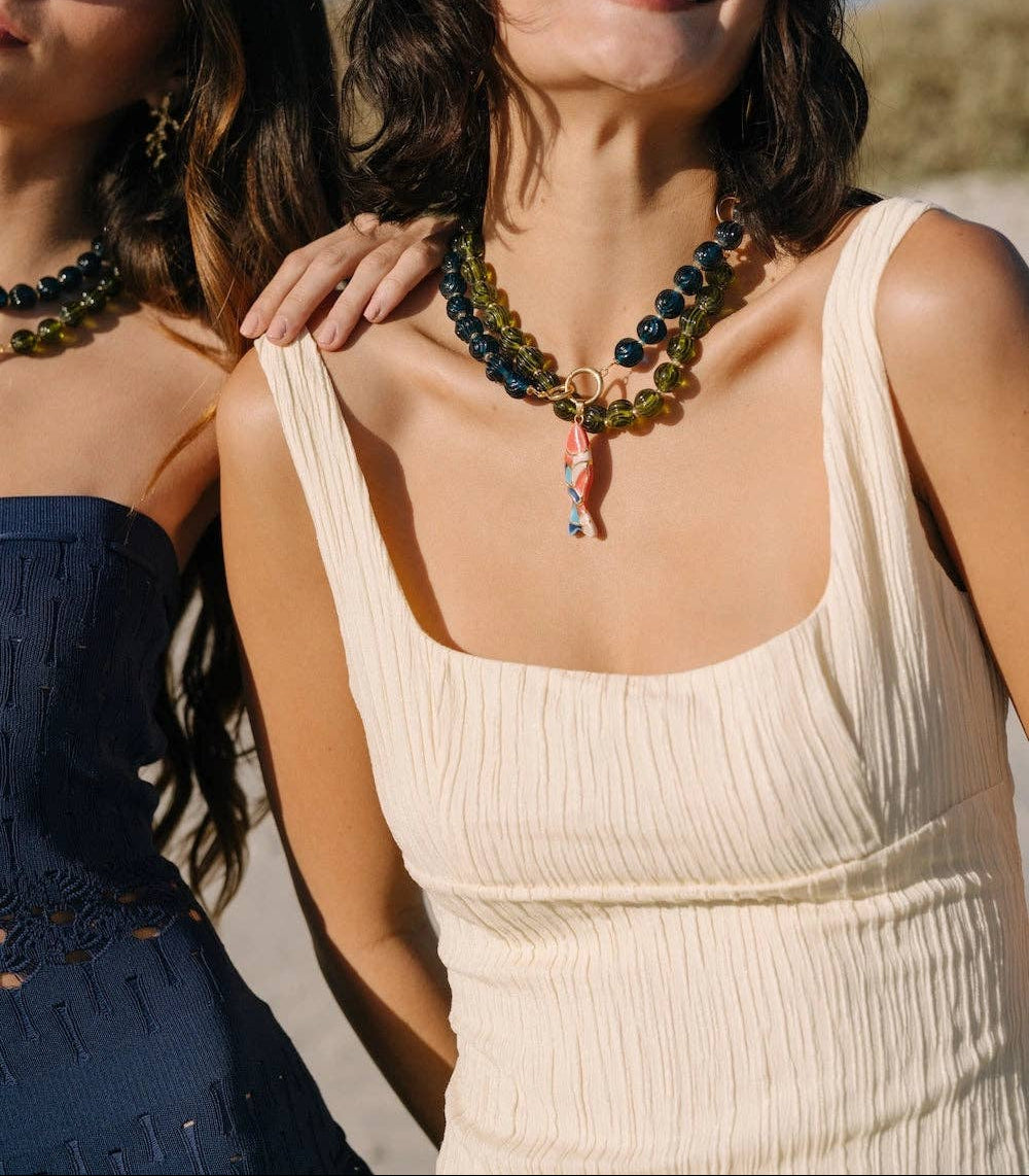 Two women outdoors wearing chunky beaded statement necklaces. The woman in the foreground wears a cream ribbed tank top and a dark green and blue beaded necklace with a colorful pendant.