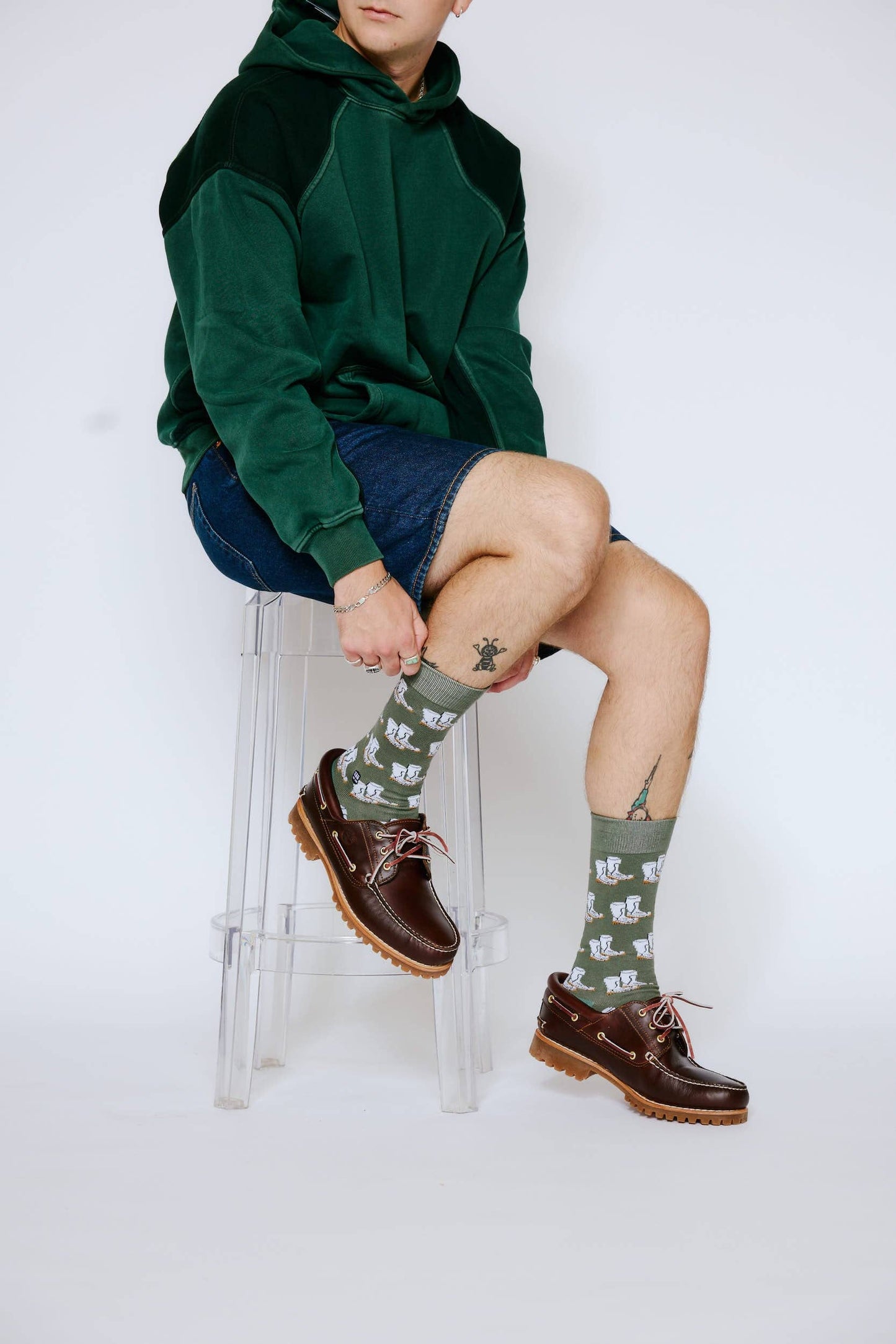 Person wearing a green hoodie and denim shorts sitting on a stool, with a white background