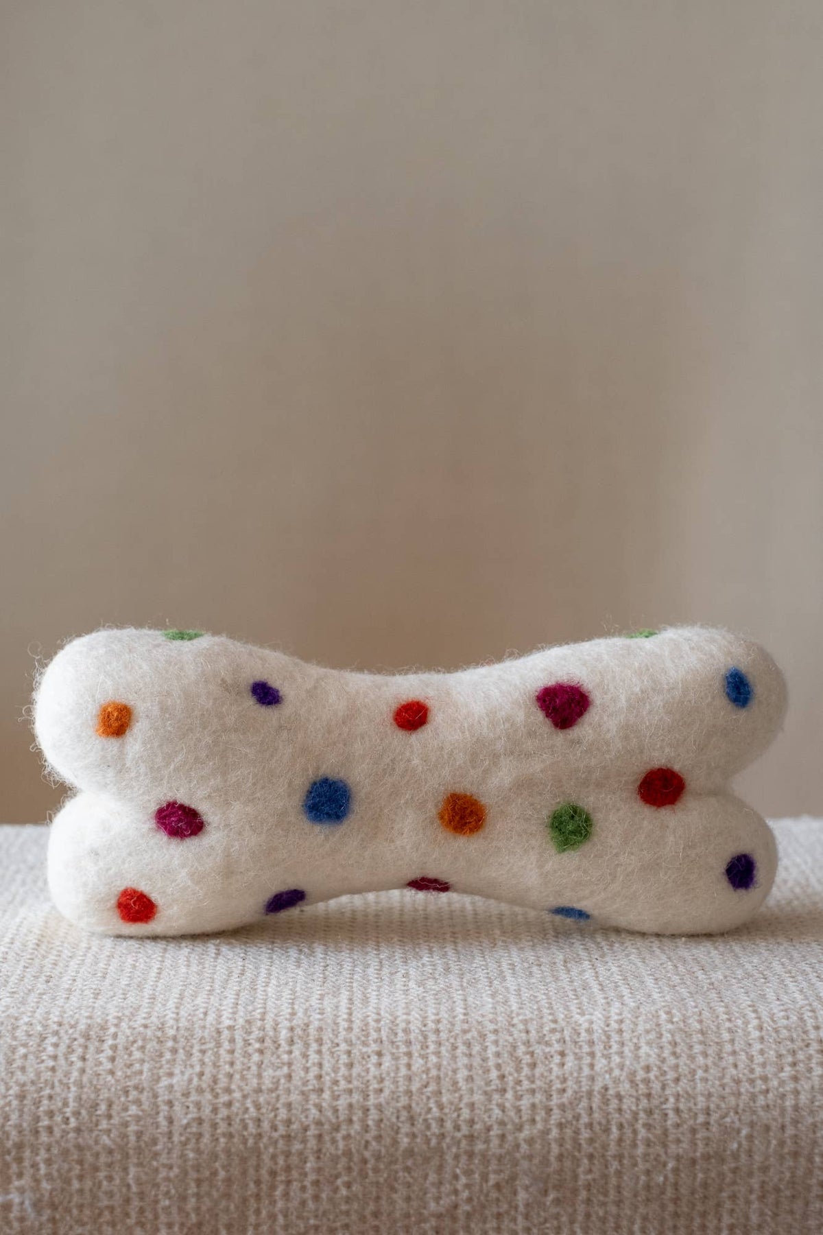 A white felted wool bone-shaped pet toy with colorful polka dots, resting on a textured cream knit blanket against a neutral beige background.