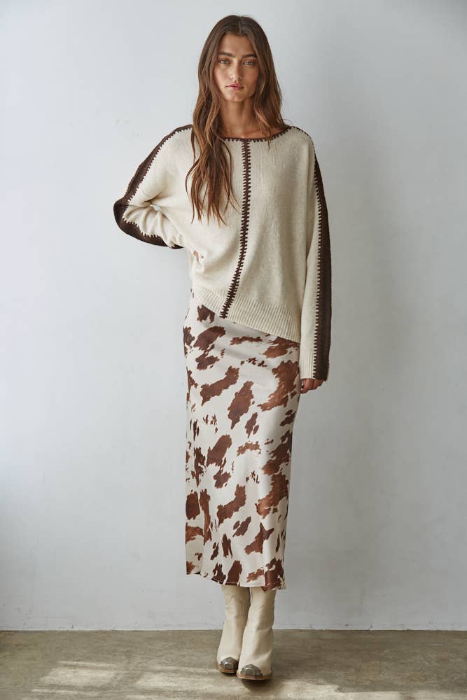 Woman wearing a cream sweater with brown accents and a cow print skirt against a plain background