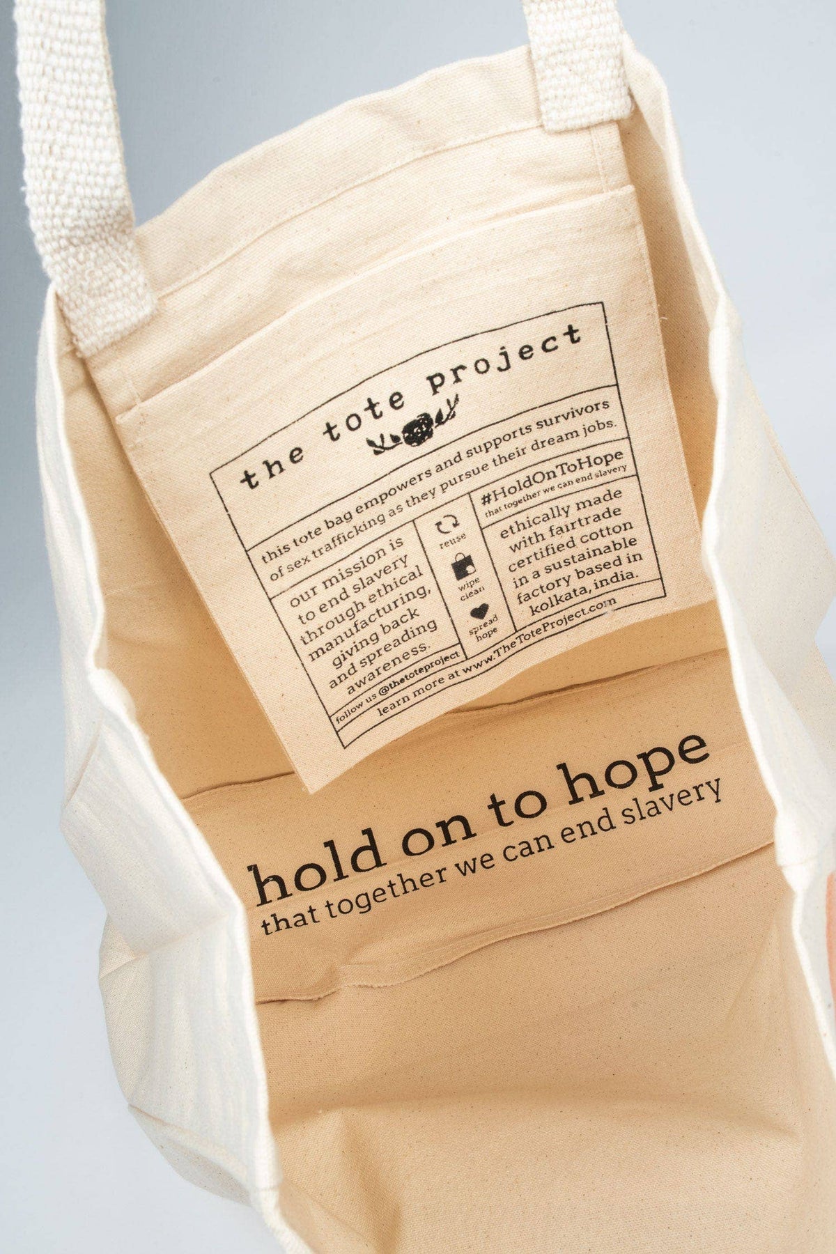 A beige canvas tote bag shown from the inside, with printed text and a paper insert. The bag has white woven handles and a warm natural canvas body.