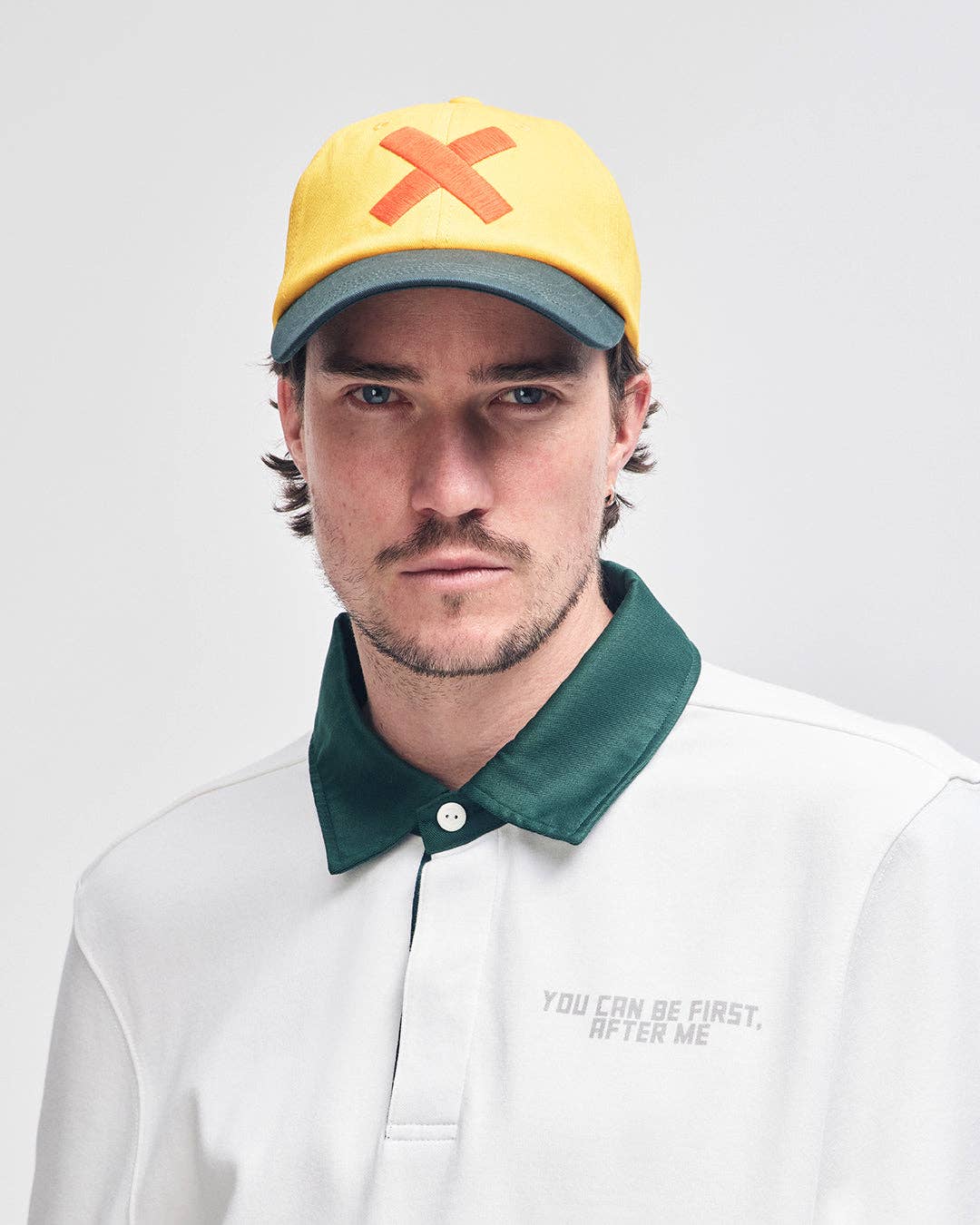 Man wearing a yellow cap with an orange 'X' and a white shirt with text on a light gray background