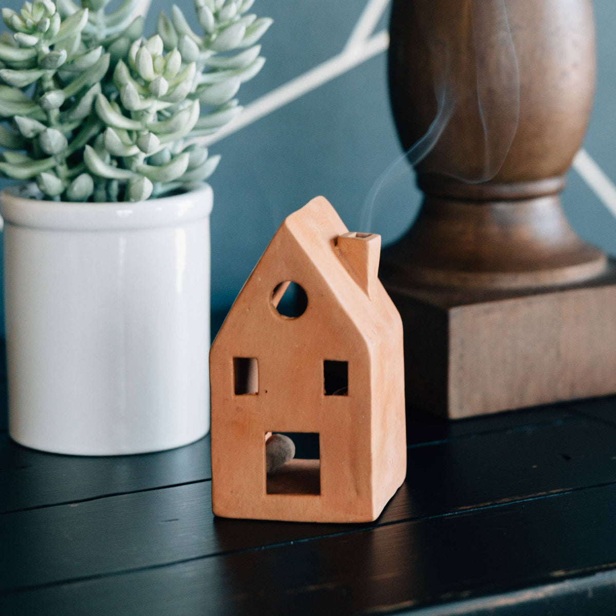 Wooden house-shaped incense burner with smoke, next to a potted plant and wooden object on a dark surface.