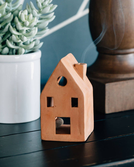 Wooden house-shaped incense burner with smoke, next to a potted plant and wooden object on a dark surface.