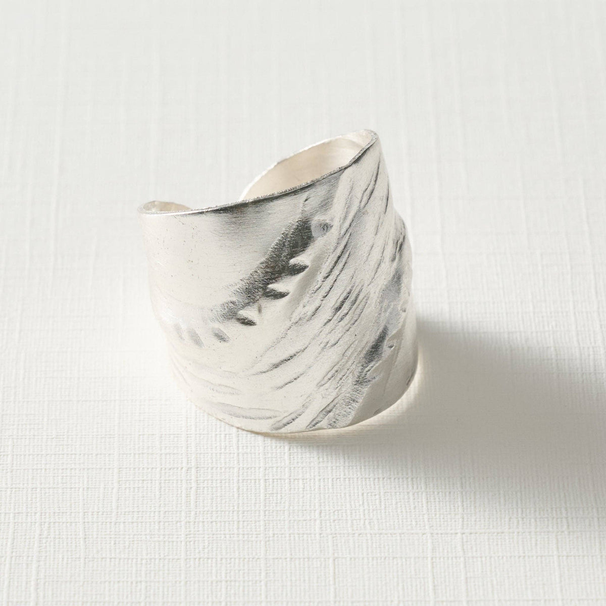 A wide, open cuff ring made of textured silver-toned metal, photographed on a light off-white fabric background.