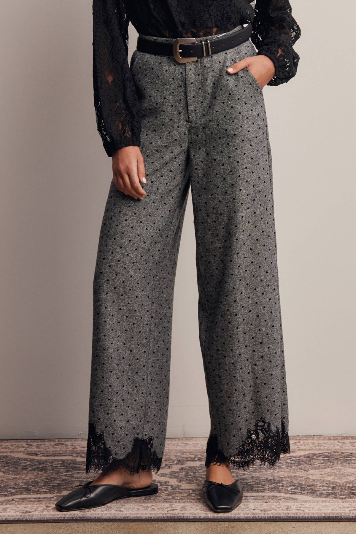 A person wearing high-waisted wide-leg gray pants with a black allover print and black lace trim at the hems, styled with a black belt, black top, and black flats against a neutral studio backdrop.