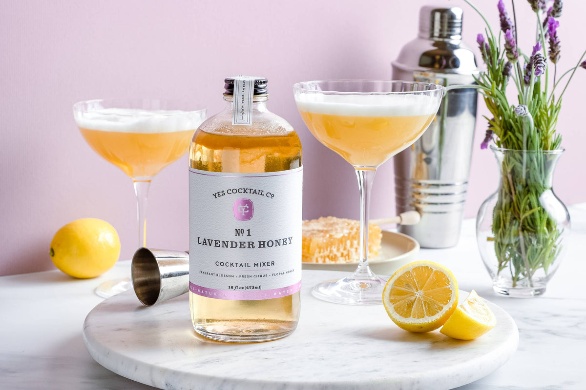 Bottle of Lavender Honey cocktail mixer with two cocktails and lemon slices on a marble surface.