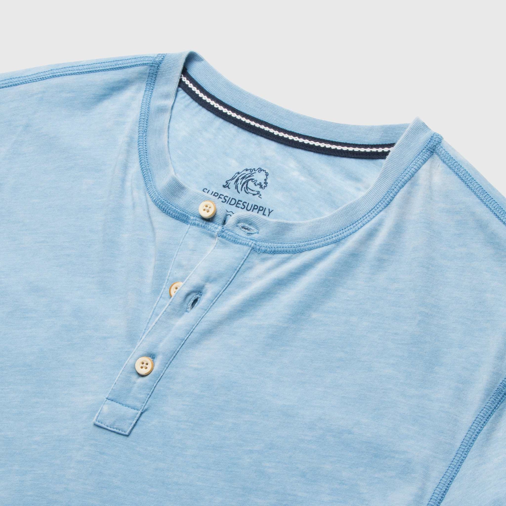 Close-up of a light blue henley shirt with buttons and a brand logo on a light gray background.