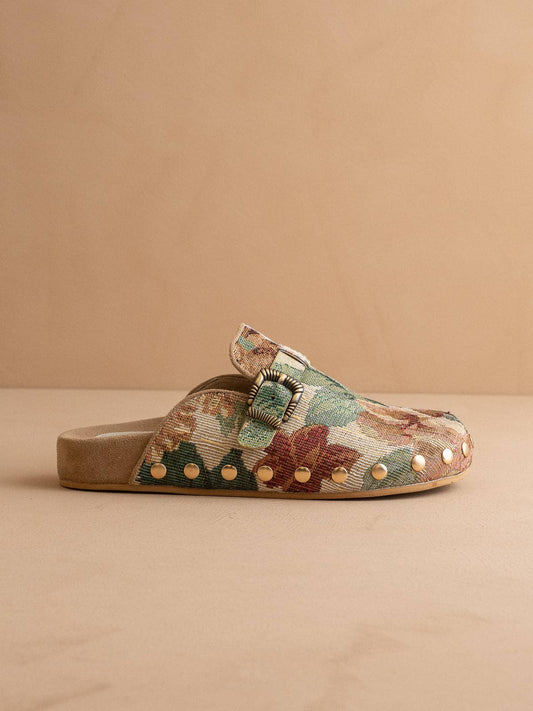 A single slip-on mule sandal with a woven floral pattern in muted green, terracotta, and beige, featuring gold-tone metal studs along the side and a tan sole, set against a solid beige background.