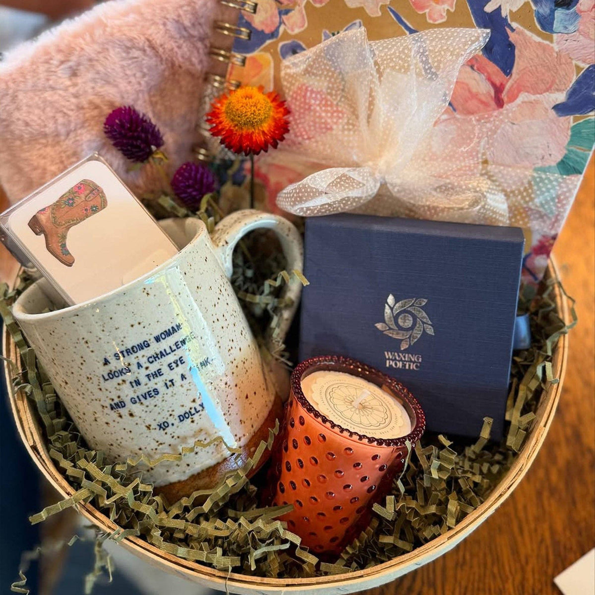 A gift basket arrangement featuring a speckled cream ceramic mug with printed text, a small orange candle, a navy blue box with a silver logo, shredded paper filler, dried flowers, and a fuzzy pink plush item in a woven basket.