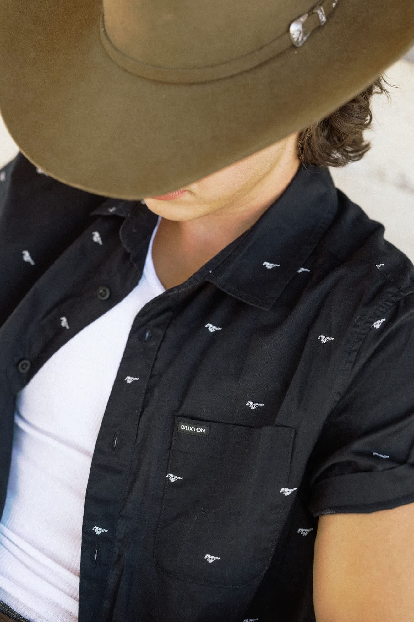 A close-up of a person wearing a black short-sleeve button-up shirt with a small white western-style logo pattern, layered over a white t-shirt and a brown cowboy hat.