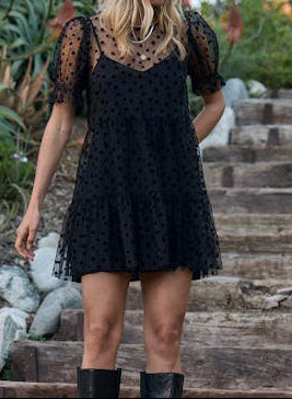 Woman wearing a black polka dot dress standing on stone steps with greenery in the background