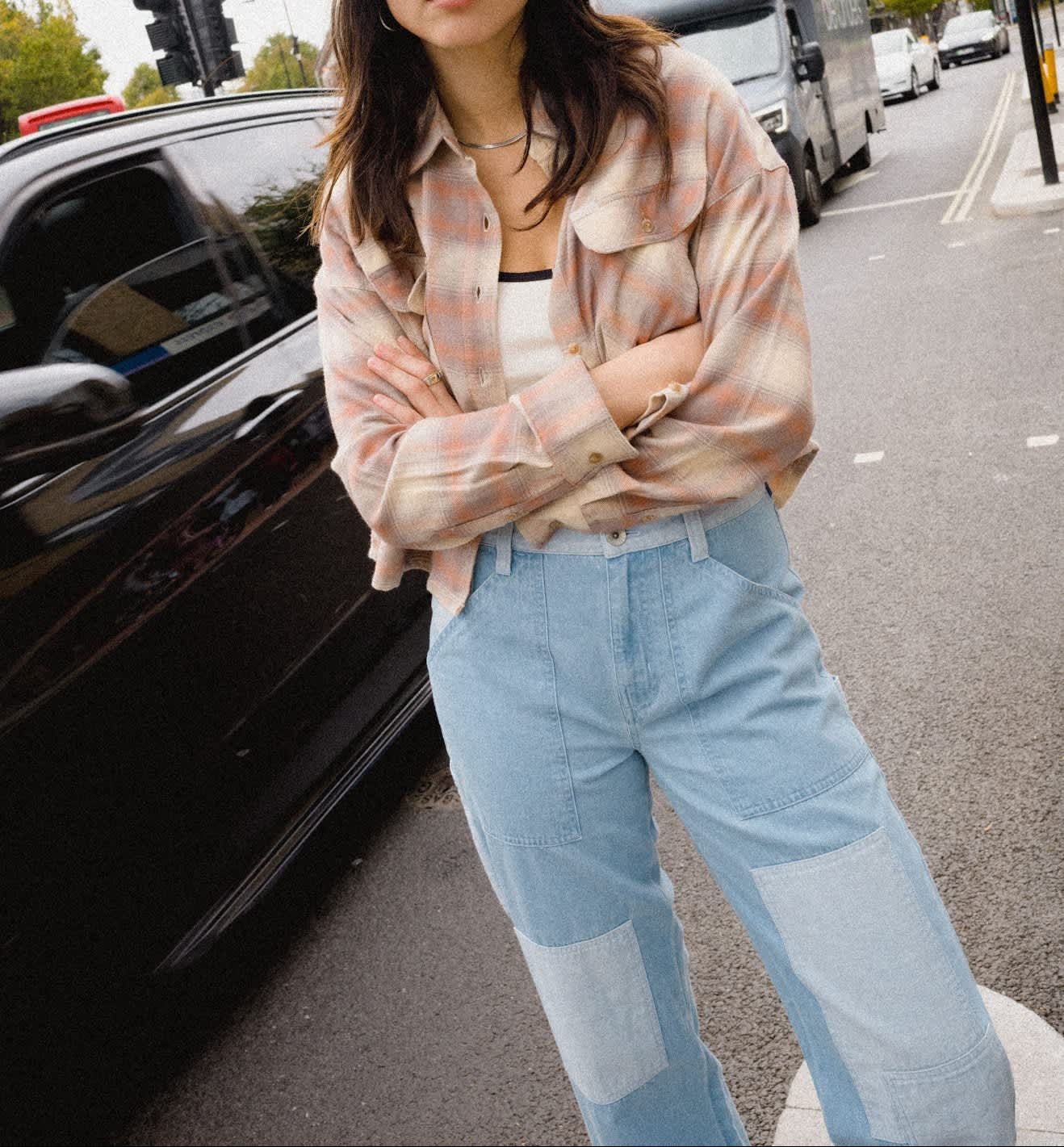 A woman standing on a city street wearing a wide-brimmed cream cowboy hat, a light pink and white plaid button-up shirt, a white top underneath, and light blue patchwork jeans.