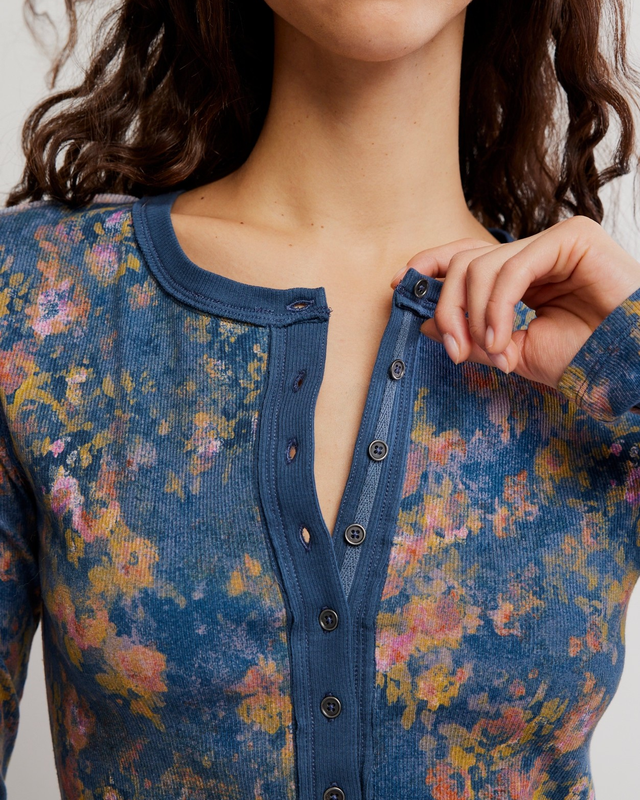Close-up of a woman wearing a long-sleeve button-front top with a blue base and multicolor floral print. She is adjusting the front closure with one hand.