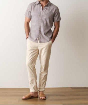 Man wearing a gray checkered shirt and beige pants standing against a plain white wall.