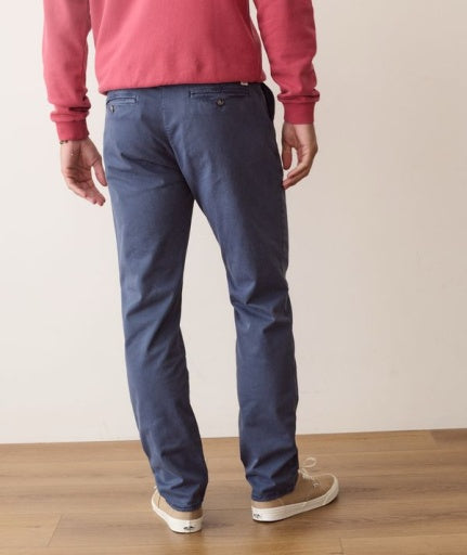 A person shown from the back wearing blue slim-fit chino pants, a pink sweatshirt, and beige sneakers, standing on a light wood floor against a plain off-white wall.