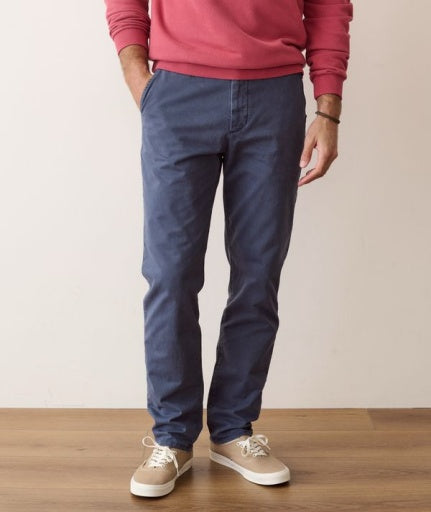 A person wearing slim-fit blue chinos with a pink sweatshirt and beige sneakers, standing against a plain beige wall and wooden floor.