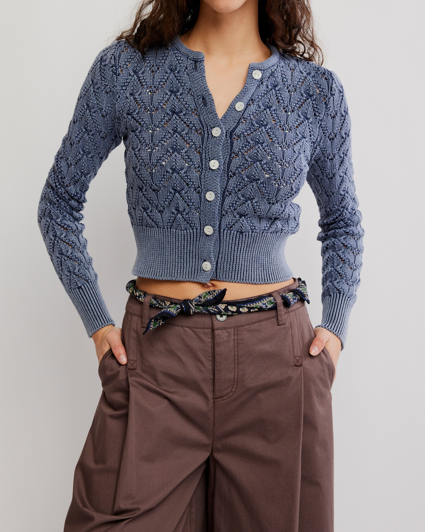 A model wearing a blue cable-knit cropped cardigan with white buttons, paired with brown wide-leg trousers and a patterned belt, against a light gray studio background.