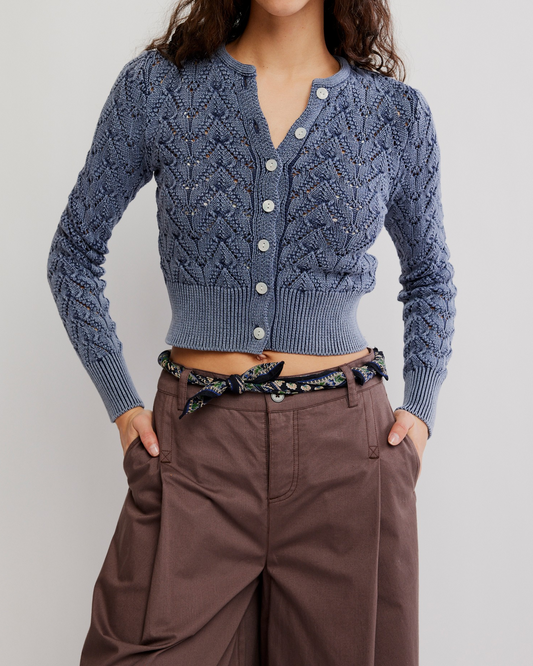 A model wearing a blue cable-knit cropped cardigan with white buttons, paired with brown wide-leg trousers and a patterned belt, against a light gray studio background.