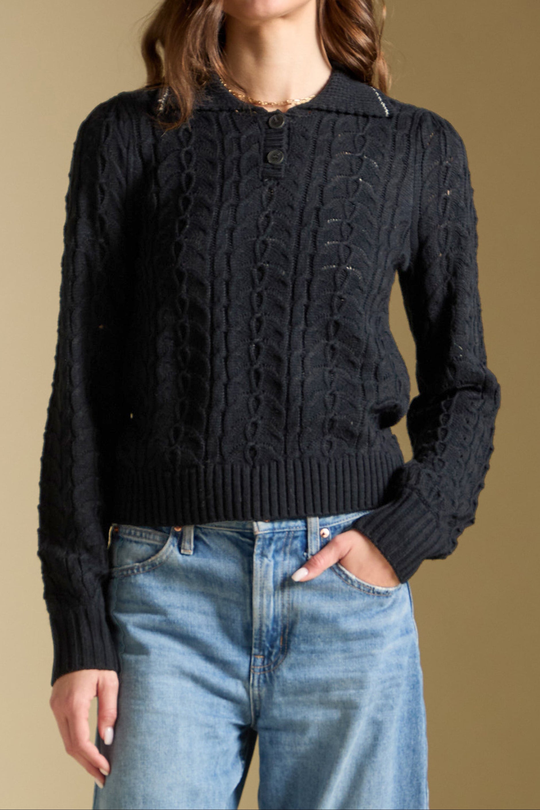 Woman wearing a black cable knit sweater and blue jeans against a beige background