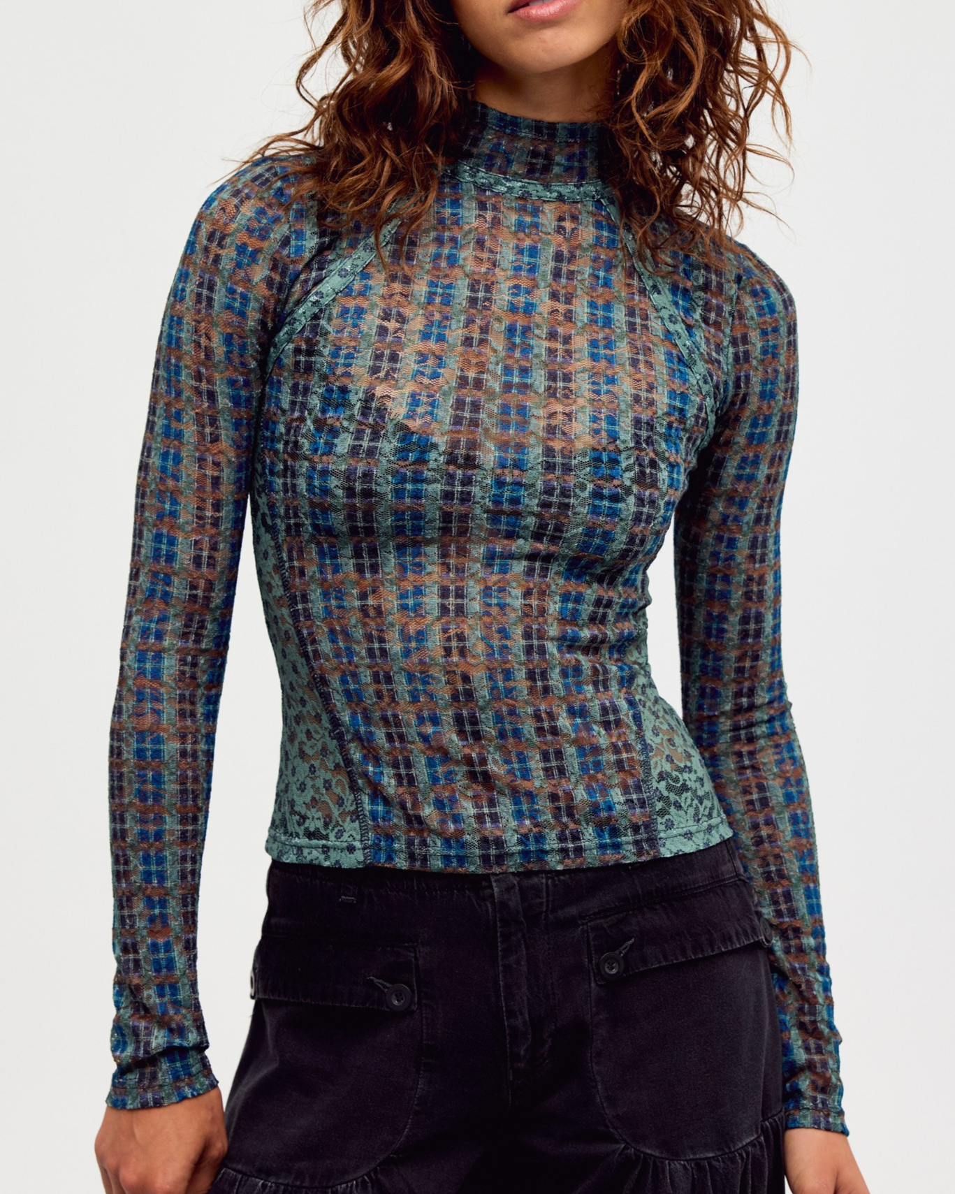 A woman wearing a long-sleeve mock neck top with a blue, teal, and brown plaid pattern, paired with dark cargo pants, against a plain light gray background.