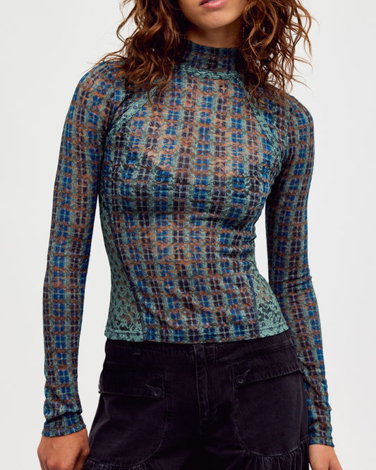 A woman wearing a long-sleeve mock neck top with a blue, teal, and brown plaid pattern, paired with dark cargo pants, against a plain light gray background.