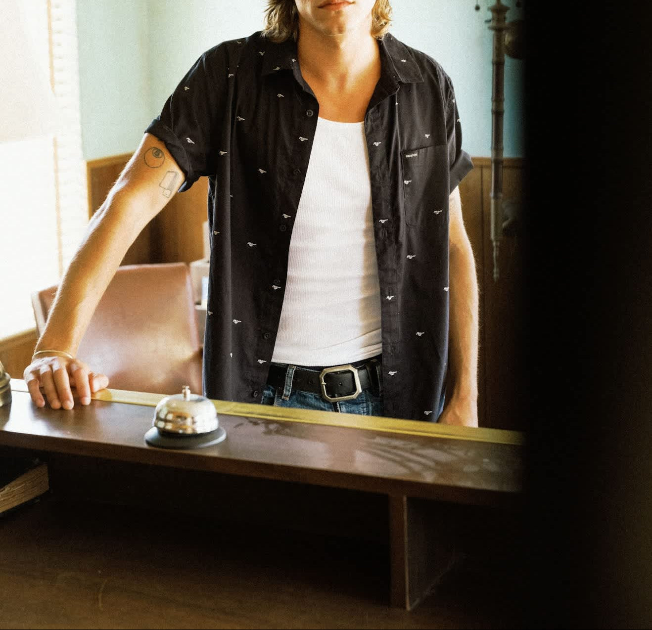 A man with long light brown hair stands indoors behind a wooden counter, wearing a black short-sleeve button-up shirt with small white embroidered details over a white tank top and blue jeans.