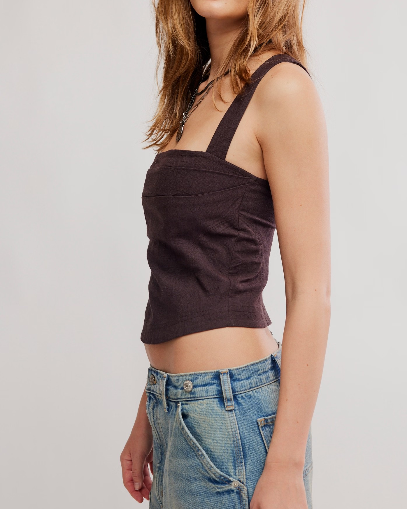 A woman wearing a dark brown cropped corset top with wide shoulder straps and blue jeans, shown from the side against a light gray studio background.