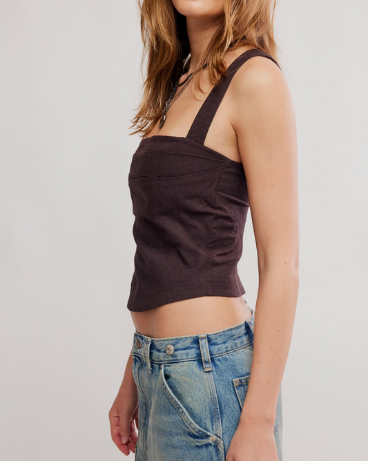 A woman wearing a dark brown cropped corset top with wide shoulder straps and blue jeans, shown from the side against a light gray studio background.
