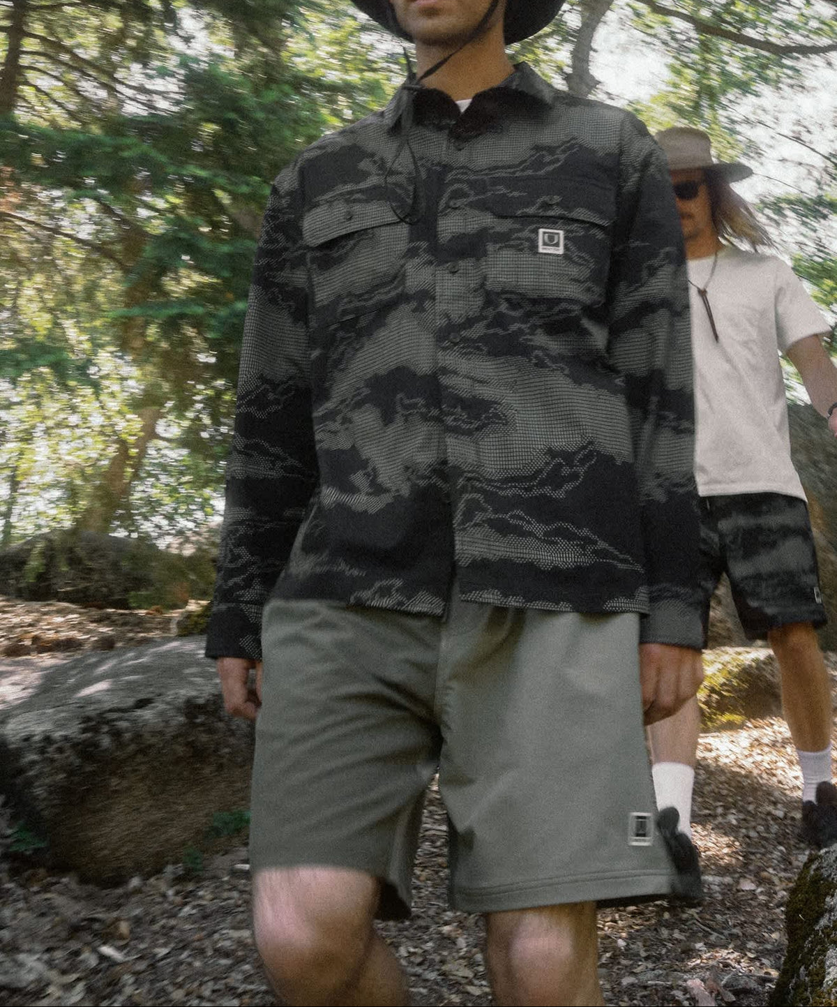 A man wearing a dark gray and black camouflage long-sleeve button-up shirt, olive green shorts, and a black bucket hat stands in a wooded outdoor setting. Another person is visible in the background.
