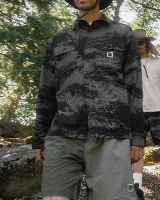 A man wearing a dark gray and black camouflage long-sleeve button-up shirt, olive green shorts, and a black bucket hat stands in a wooded outdoor setting. Another person is visible in the background.