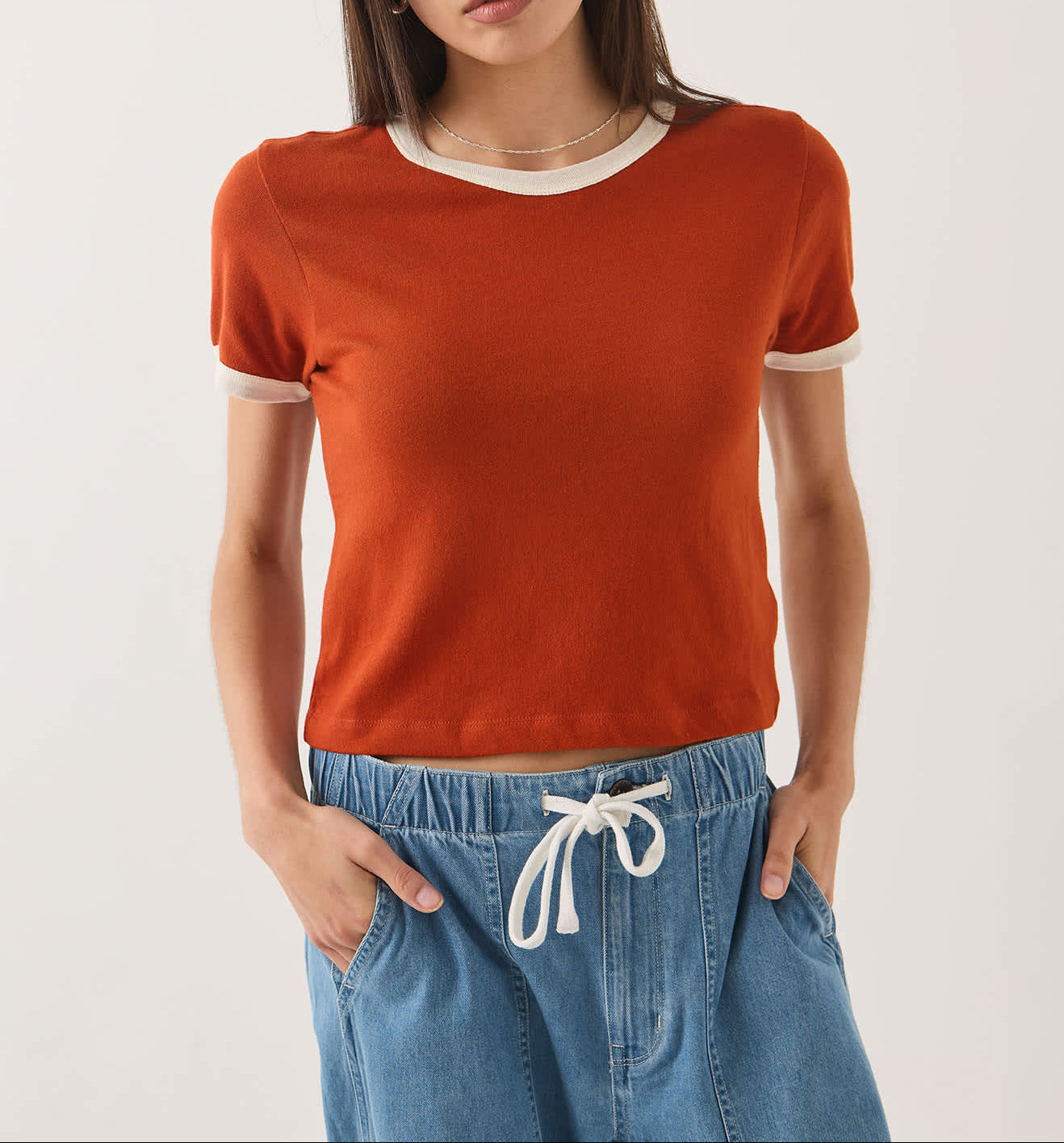 A woman modeling a short-sleeve crewneck knit top in burnt orange with contrasting white trim at the collar and sleeve cuffs, paired with light blue jeans against a plain light gray studio background.