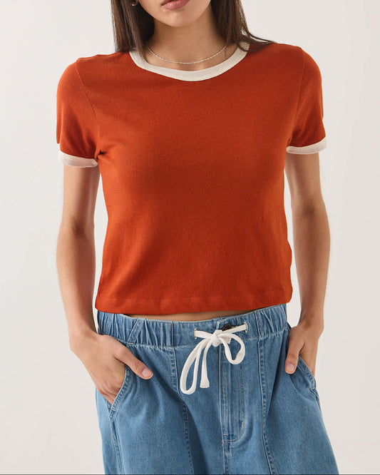 A woman modeling a short-sleeve crewneck knit top in burnt orange with contrasting white trim at the collar and sleeve cuffs, paired with light blue jeans against a plain light gray studio background.
